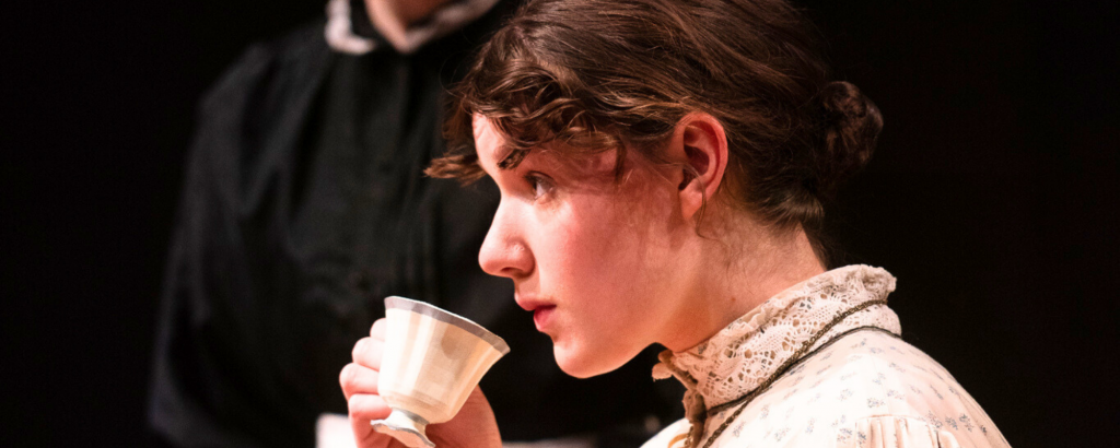 Emilie (Morgan Hrymack) hesitantly sipping tea as she attempts to make sense of her new environment in Connecticut Rep's production of The Moors (Fall 2024).