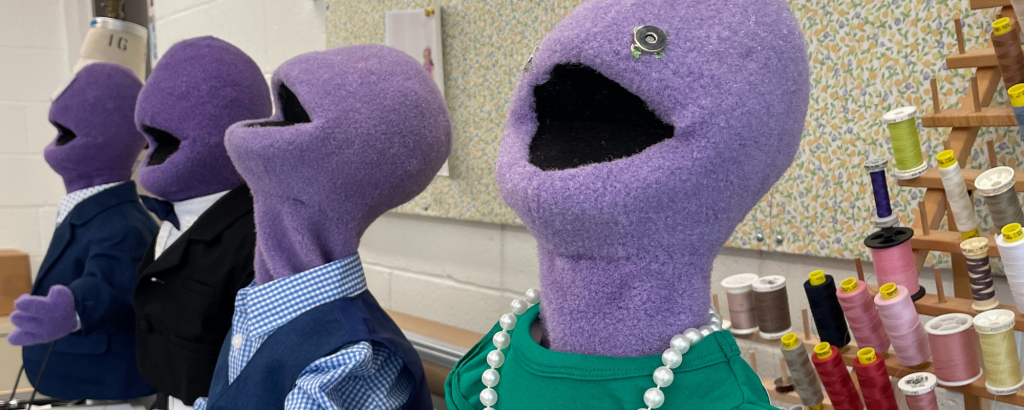 Four puppets covered in purple felt in a row in varying levels of professional attire in front of a collection of different colored threads for Connecticut Rep's War of the Worlds (Fall 2023).