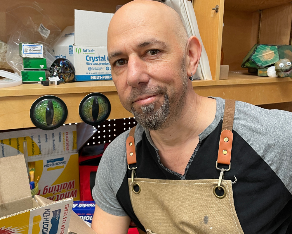 Portrait of Jaron Hollander, a white man with facial hair wearing a heavy duty apron in the puppet shop.