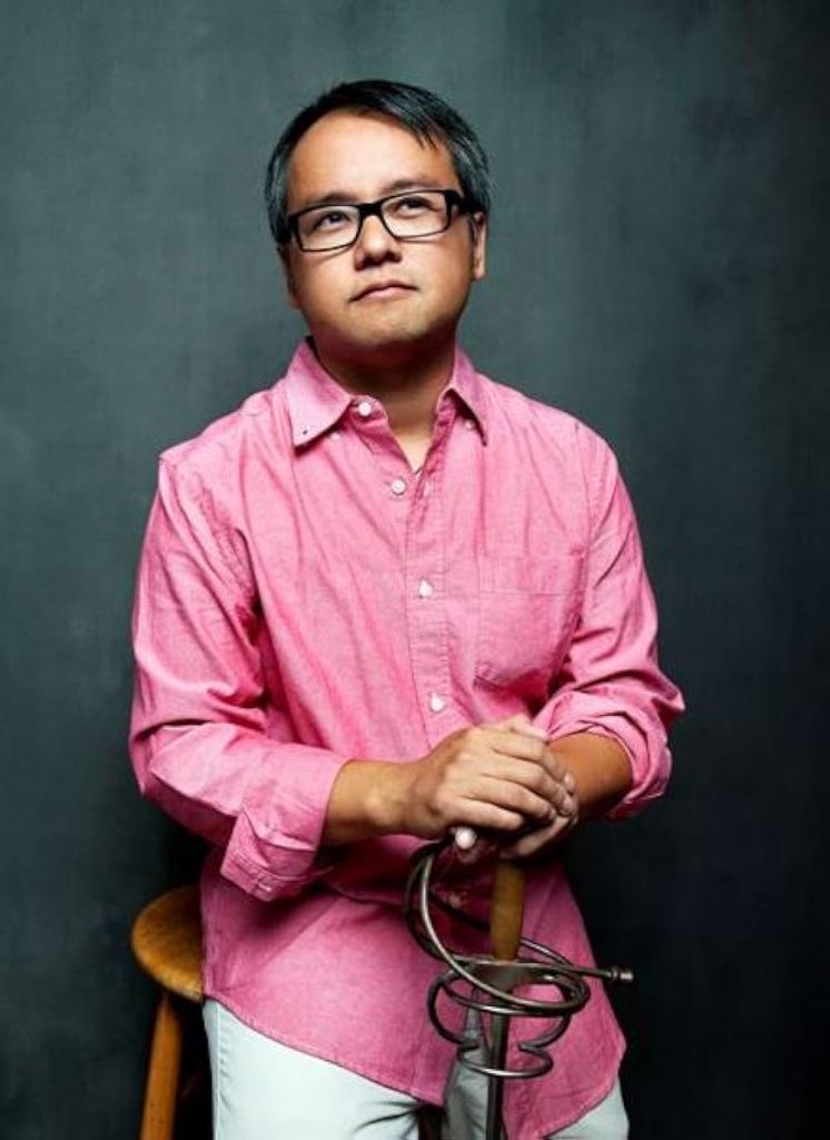 Portrait of playwright Qui Nguyen, an Asian man with black glasses, wearing a pink button-down shirt and nonchalantly holding a sword.