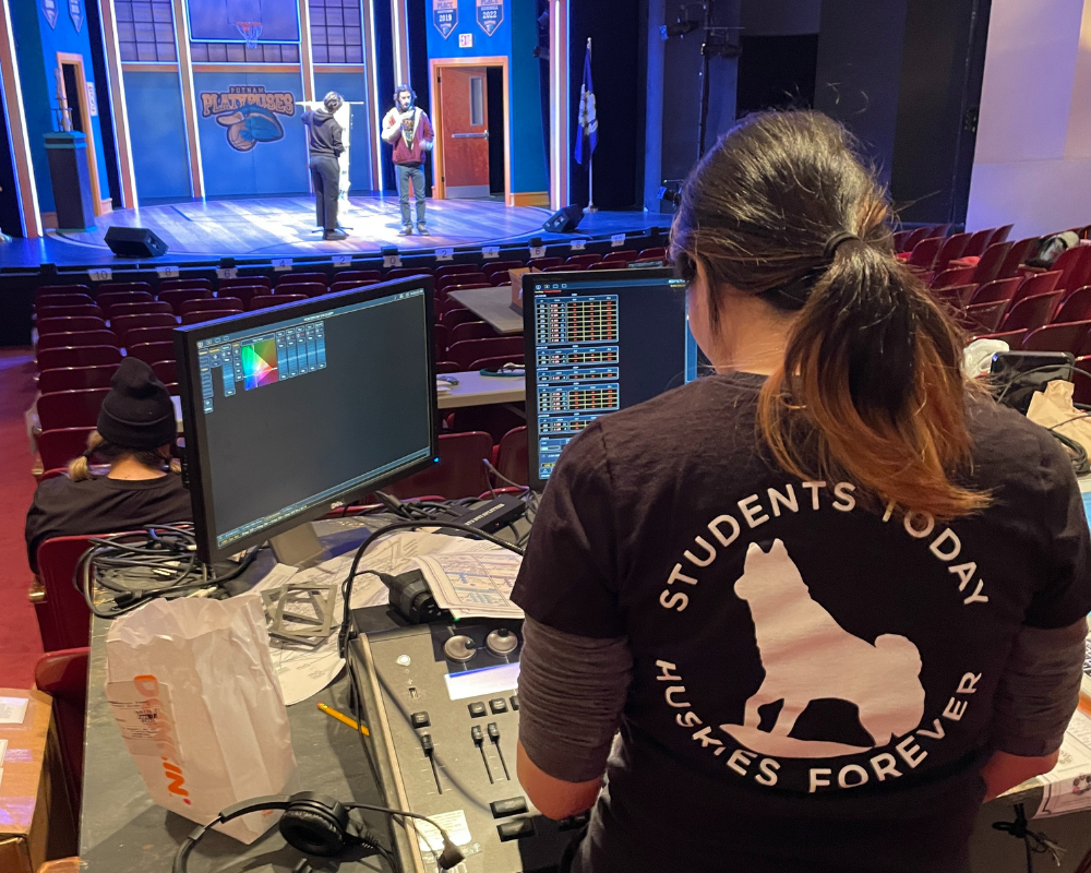 A student sits at a bank of monitors and works the light board during a technical rehearsal of The 23rd Annual Putnam County Spelling Bee.