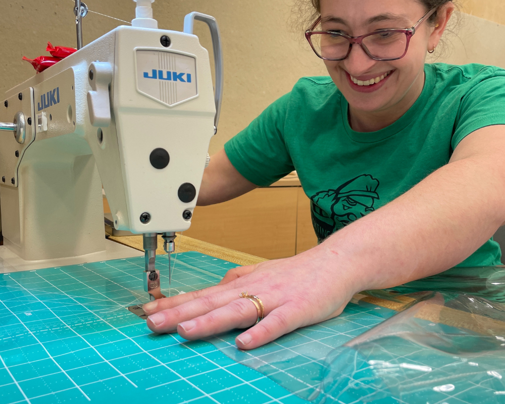A smiling student sews sheets of clear vinyl.