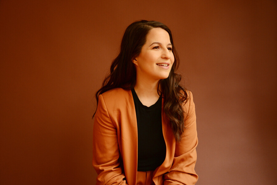 Portrait of songwriter Shaina Taub, a young woman with long dark hair, wearing an orange jacket, looking away from the camera.