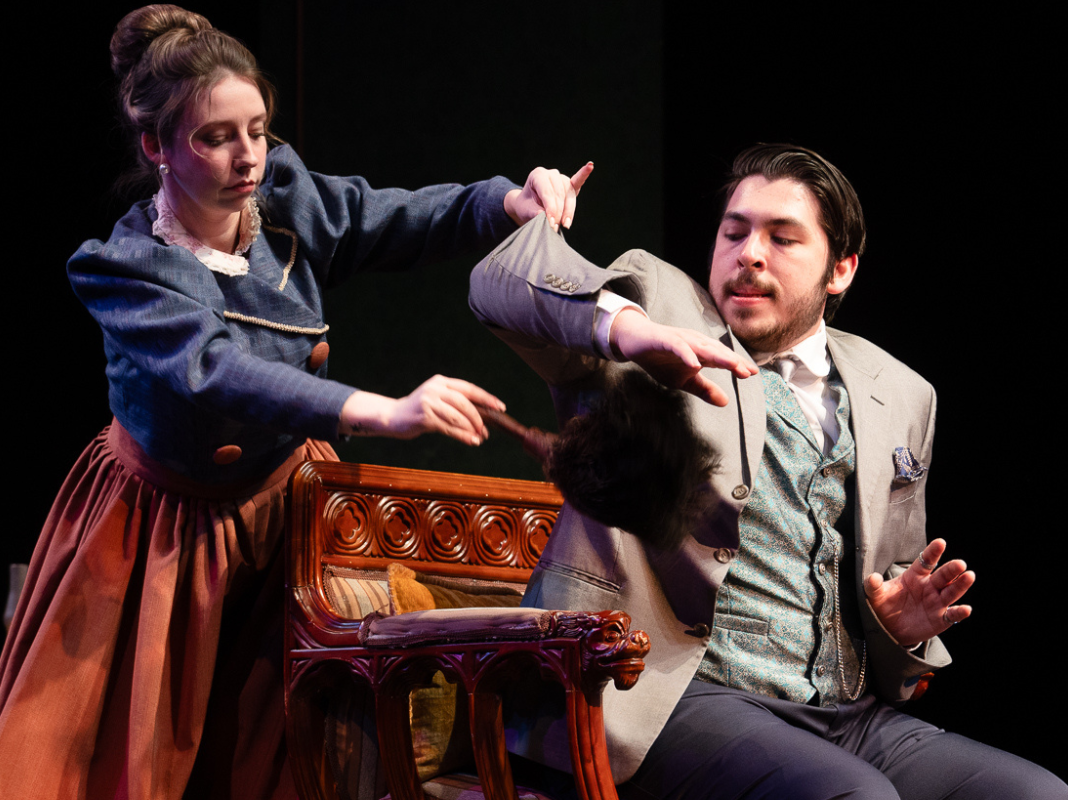 An ambitious maid dusts under the arm of a serious gentleman in the 2024 production of A Doctor's Dilemma.