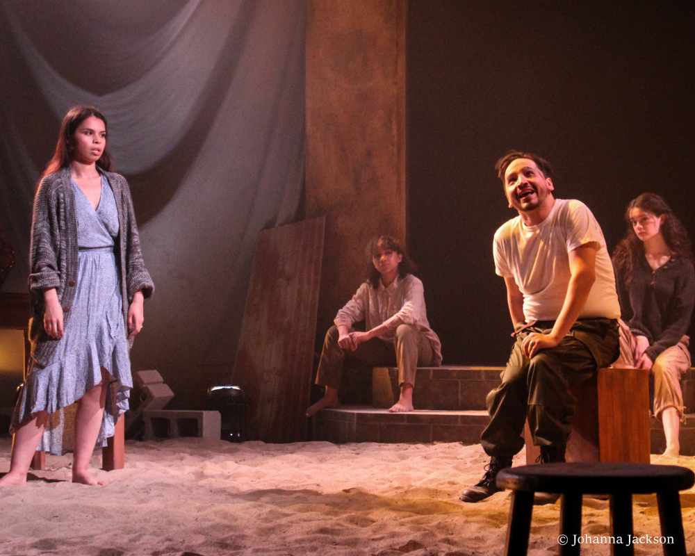 A young woman in a flowing blue dress l listens to a young man in t-shirt and jeans, sitting on a stool, in the CRT production of Seven Spots on the Sun.