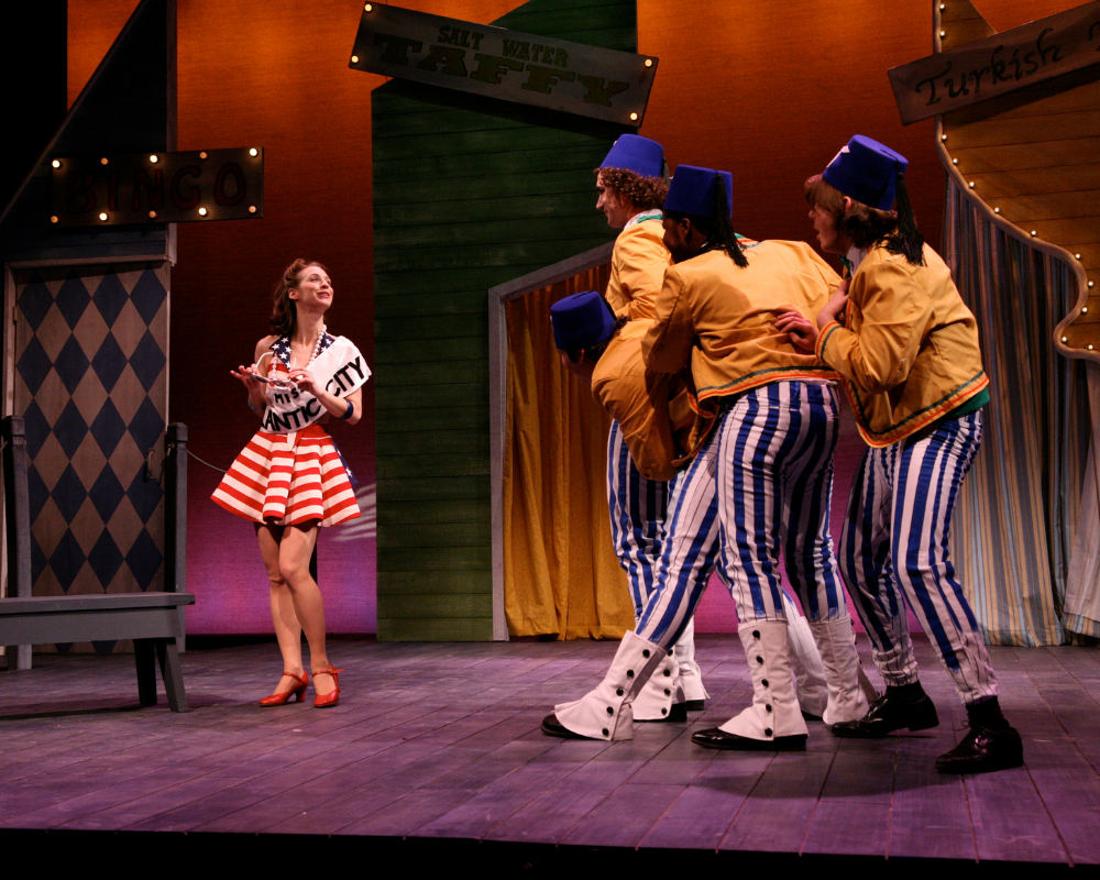 Dressed in a short red, white and blue dress and wearing a sash reading "Miss Atlantic City," a young woman poses while three young men in yellow jackets, blue and white striped pants and blue Shriner's hats observe. The Skin of Our Teeth, 2008.