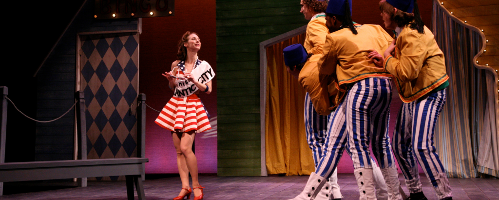 Dressed in a short red, white and blue dress and wearing a sash reading "Miss Atlantic City," a young woman poses while three young men in yellow jackets, blue and white striped pants and blue Shriner's hats observe. The Skin of Our Teeth, 2008.