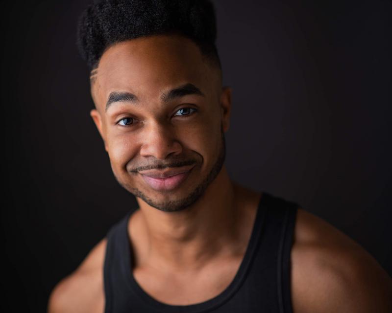 Portrait of Brandon Lamar Kelly, a Black man with short facial hair, smiling at the camera, wearing a dark top.