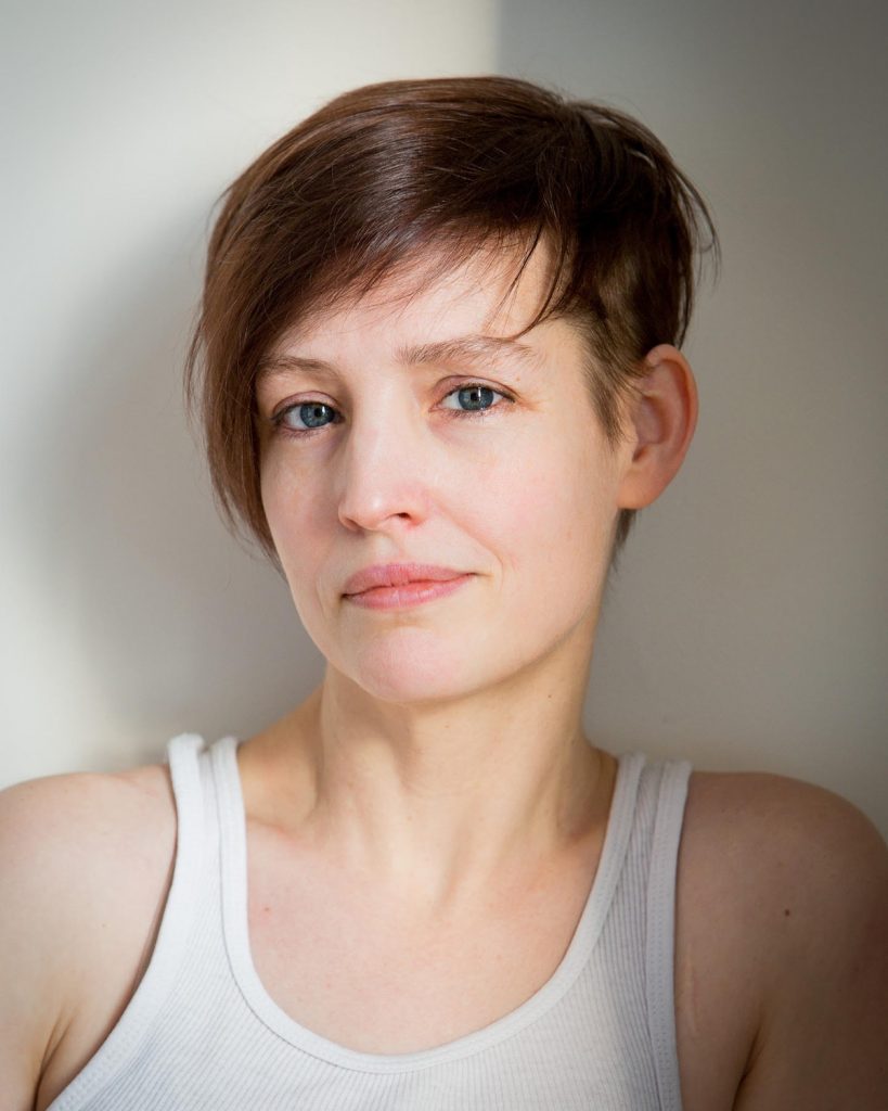 Portrait of Kate Brehm, a white woman with short hair brown hair and blue eyes, wearing a white tank top.