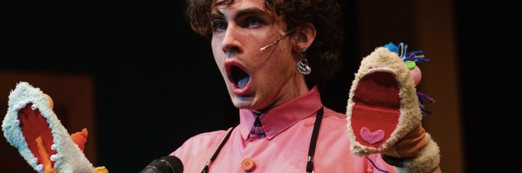 Leaf Coneybear (Aidan McNamara) from Connecticut Rep's production of The 25th Annual Putnam County Spelling Bee (Spring 2024) bellowing out his word in front of a microphone with puppets on both hands.