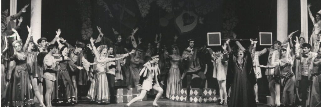 The cast of Once Upon A Mattress sing and gesture toward a woman in black holding a bar above her head.