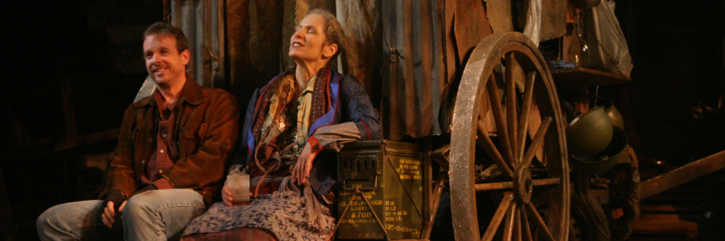 Connecticut Repertory Theatre's Mother Courage and Her Children (2004) with two actors in brown and green disheveled clothing sitting and smiling on a wagon, filled with all of their belongings.