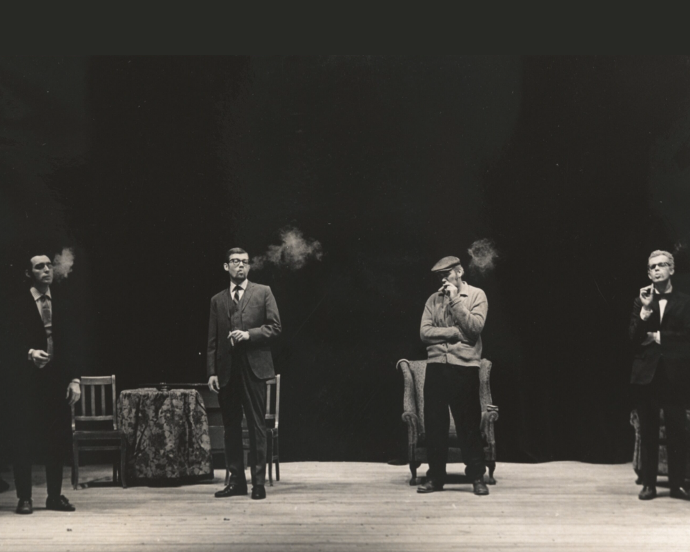 Four men from varying backgrounds, spaced equidistantly are facing the audience while smoking cigarettes.