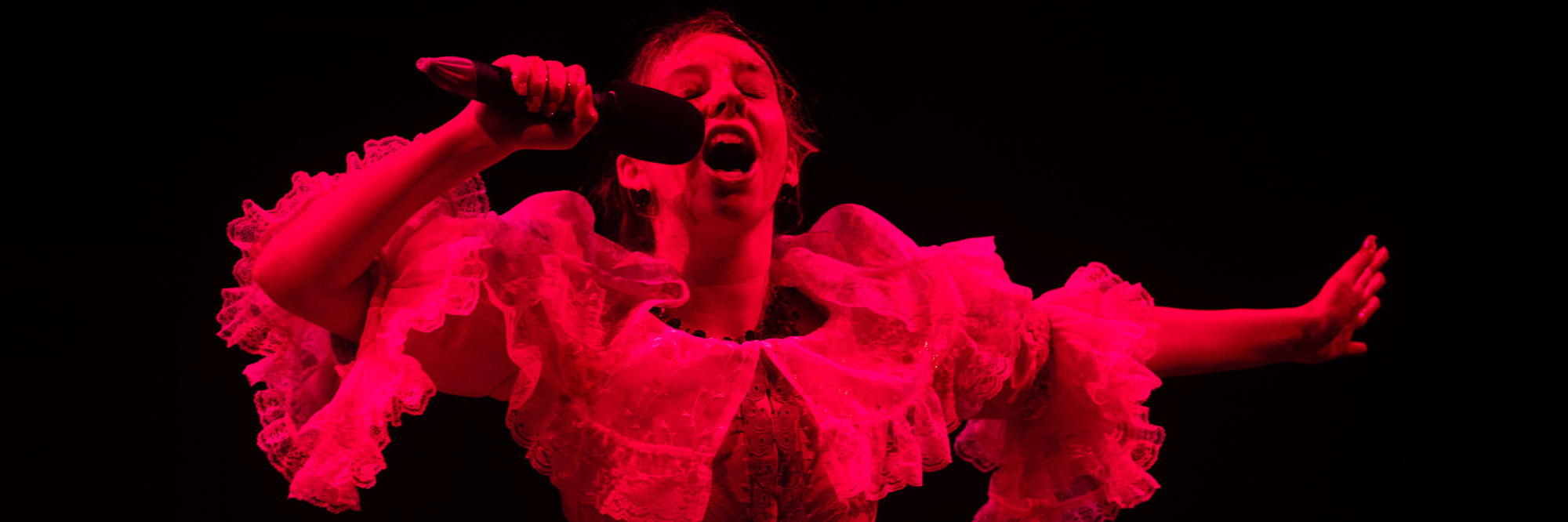 A young woman bathed in red light sings wildly into a microphone in the 2024 production of The Moors.