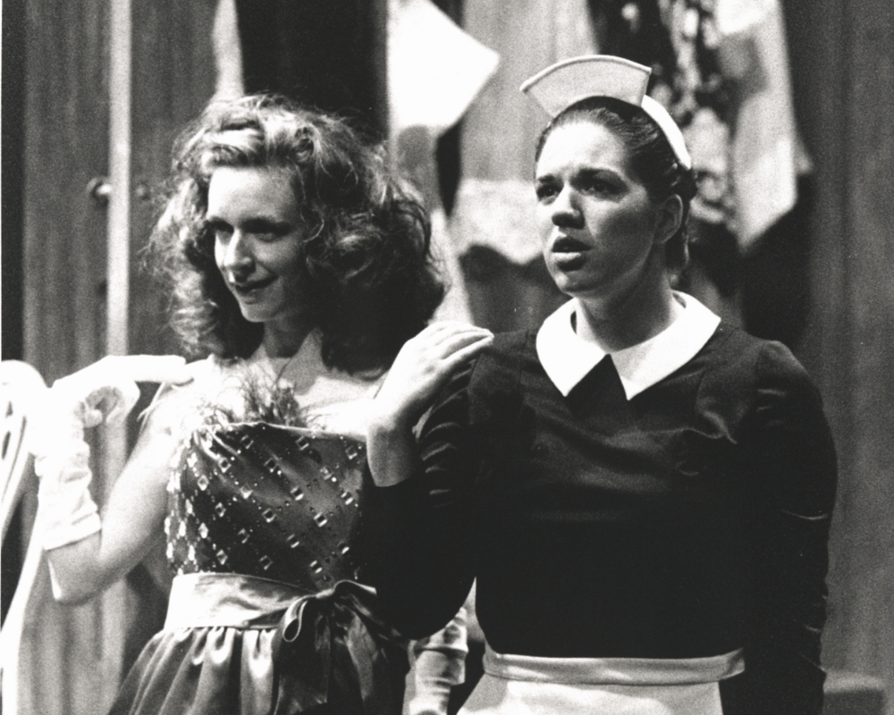 A maid stares forward as a weathy woman with brown hair flirts with someone