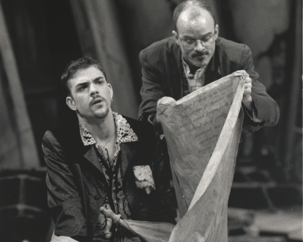 A gentleman with a thin mustache and goatee ponders while an bespectacled elderly man reads through a long scroll.