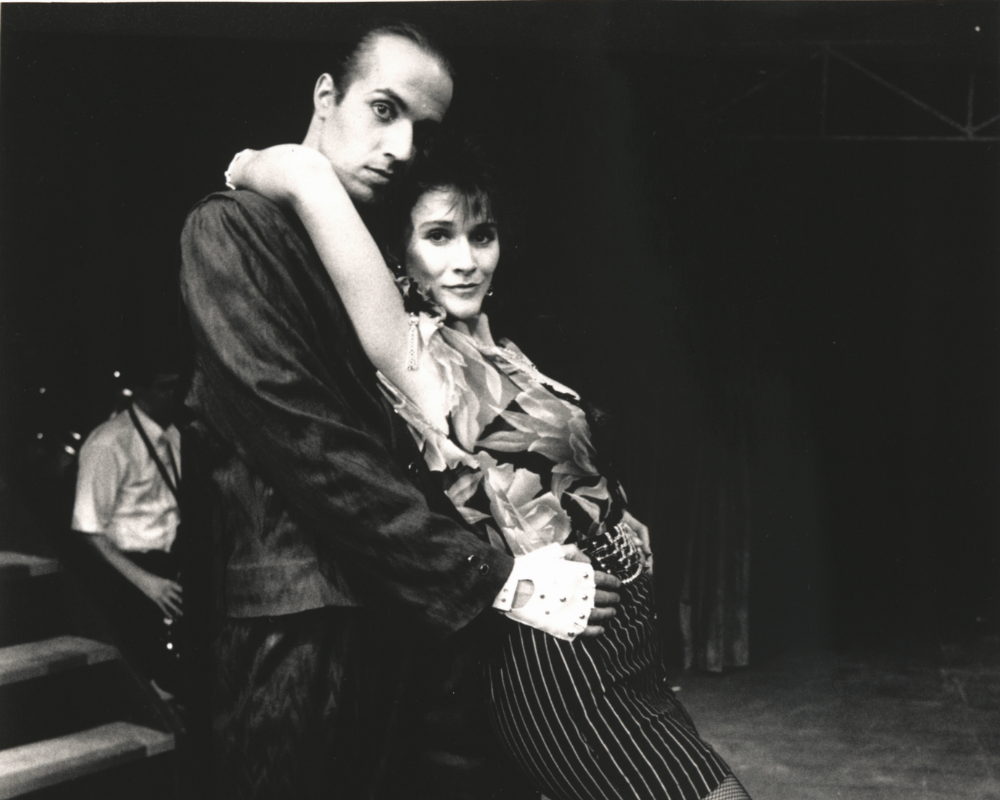 From the production of Threepenny Opera, a woman with short dark hair, wearing a striped skirt and fishnet stockings leans into a gentleman who holds her hips.